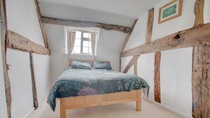 A bedroom with exposed wooden beams, a bed with floral bedding, and a window with curtains. - Beautiful 3-bed House in the Hamlet of Ham Green (Redditch)