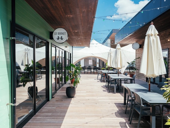 An outdoor seating area with tables and chairs, a sign reading 'LB MARKET', and a wooden ceiling.