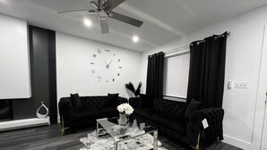 A modern living room with a black sectional sofa, a glass coffee table, a cowhide rug, a wall clock, and a ceiling fan.