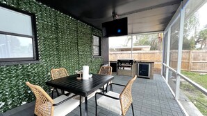 An outdoor patio with a black table and wicker chairs, a grill, and a TV mounted on the ceiling.