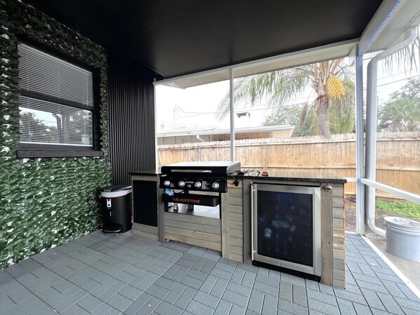 Un  área de cocina al aire libre con una parrilla, un cubo de basura y una ventana que da a un patio trasero.