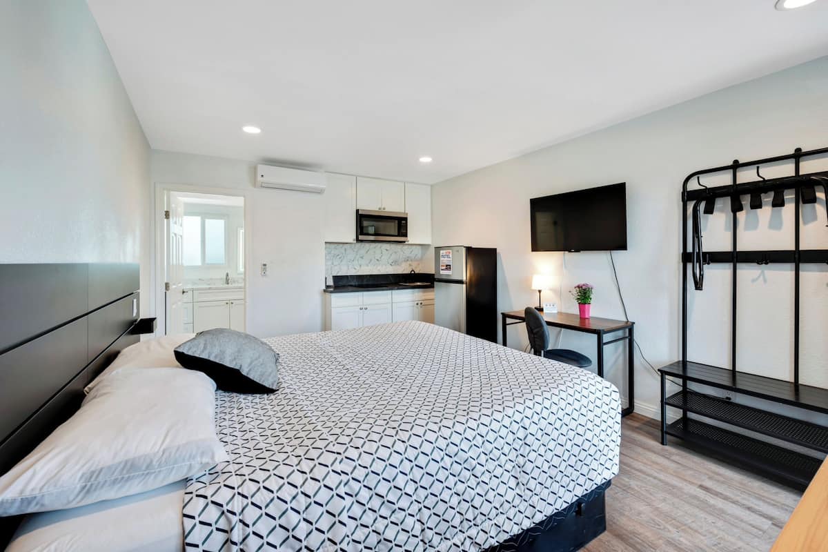 A compact hotel room with a bed, a desk, a TV, a kitchenette, and a balcony with a metal railing.