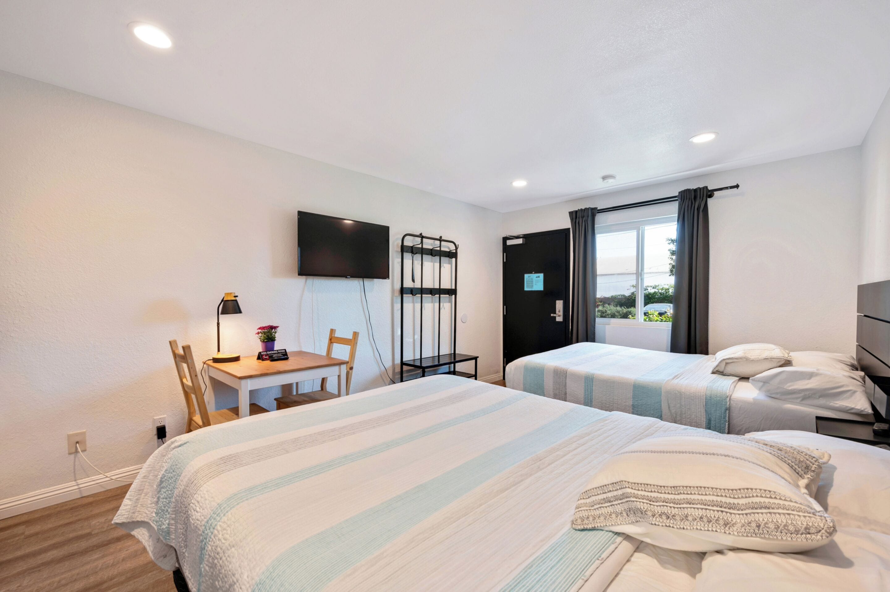 A hotel room with two beds, a desk with a chair, a TV mounted on the wall, and a window with curtains.