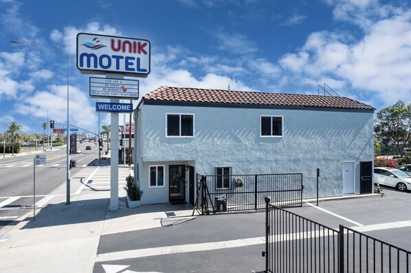 A Ulik Motel with a blue exterior and a red-tiled roof, located at a street intersection with a 'Welcome' sign. - Unik Motel (Harbor City)
