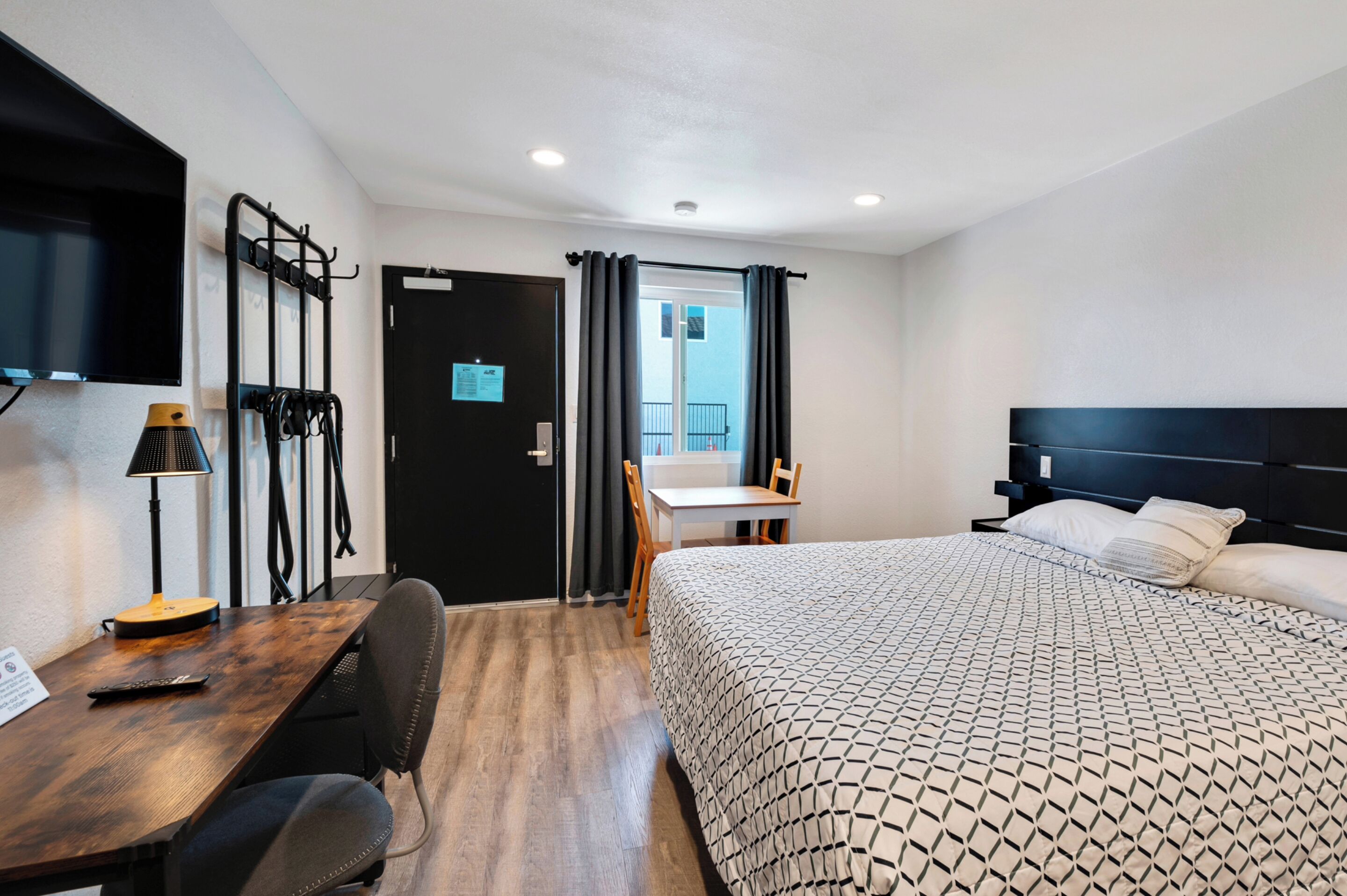 Habitación de hotel con una cama, un escritorio con lámpara, una silla, un televisor montado en la pared y una puerta que da a un balcón.