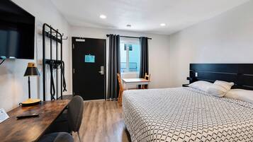 Habitación de hotel con una cama, un escritorio con lámpara, una silla, un televisor montado en la pared y una puerta que da a un balcón.