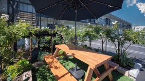 Une table de pique-nique en bois, surmontée d’un grand parasol, entourée de verdure et avec un bâtiment en arrière-plan.
