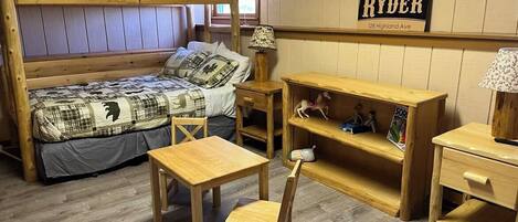 A bunk bed with a checkered bedspread, a wooden desk with a chair, a shelf with toys, and a lamp.