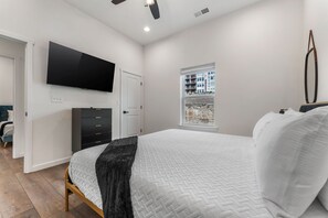 A bedroom with a bed, a TV mounted on the wall, a window with a view of buildings, and a ceiling fan. - Lakefront Rooftop Hangout Hot Tub Fire Pit Arcades (Branson)