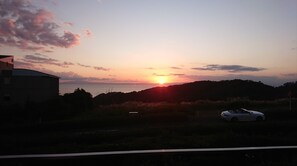 Auringonlasku kumpuilevan maiseman yllä, ja tienvarressa on pysäköitynä auto.