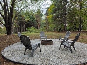 A circular seating area with Adirondack chairs and a metal fire pit surrounded by gravel, set in a forested outdoor space. - BIG Hart Lakehouse Hot Tub near Silver Lake Dunes (Hart)