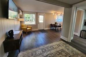 Una sala de estar moderna con piso de madera oscura, un sofá amarillo, un comedor con mesa y sillas, y un ventanal grande.