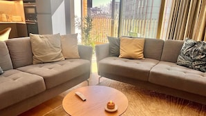Un salon moderne avec deux canapés gris, une table basse ronde en bois et une vue sur le paysage urbain, visible à travers de grandes fenêtres.