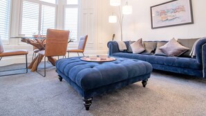 Ett vardagsrum med en blå soffa, en tillhörande pall och en matplats med ett glasbord och stolar.