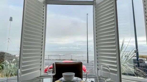 En matplats med ett bord dukat för två personer, utsikt över havet genom stora fönster och en röd bil parkerad utanför.
