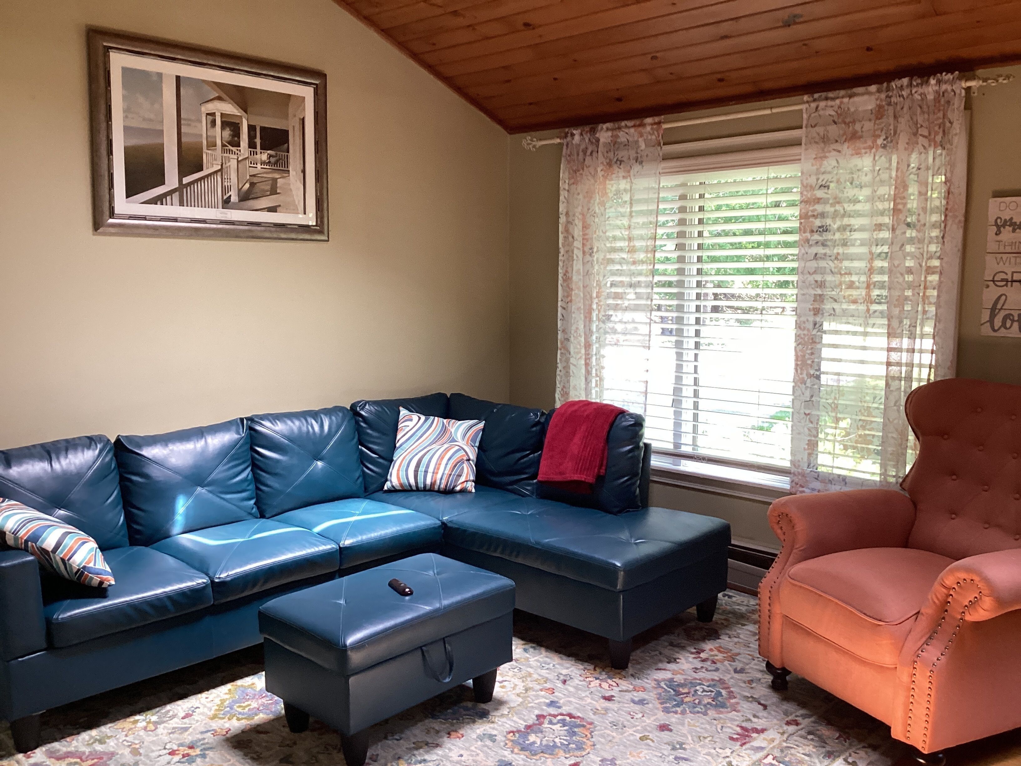 A living room with a sectional sofa, a small ottoman, a pink armchair, a window with blinds, and a framed picture on the wall.