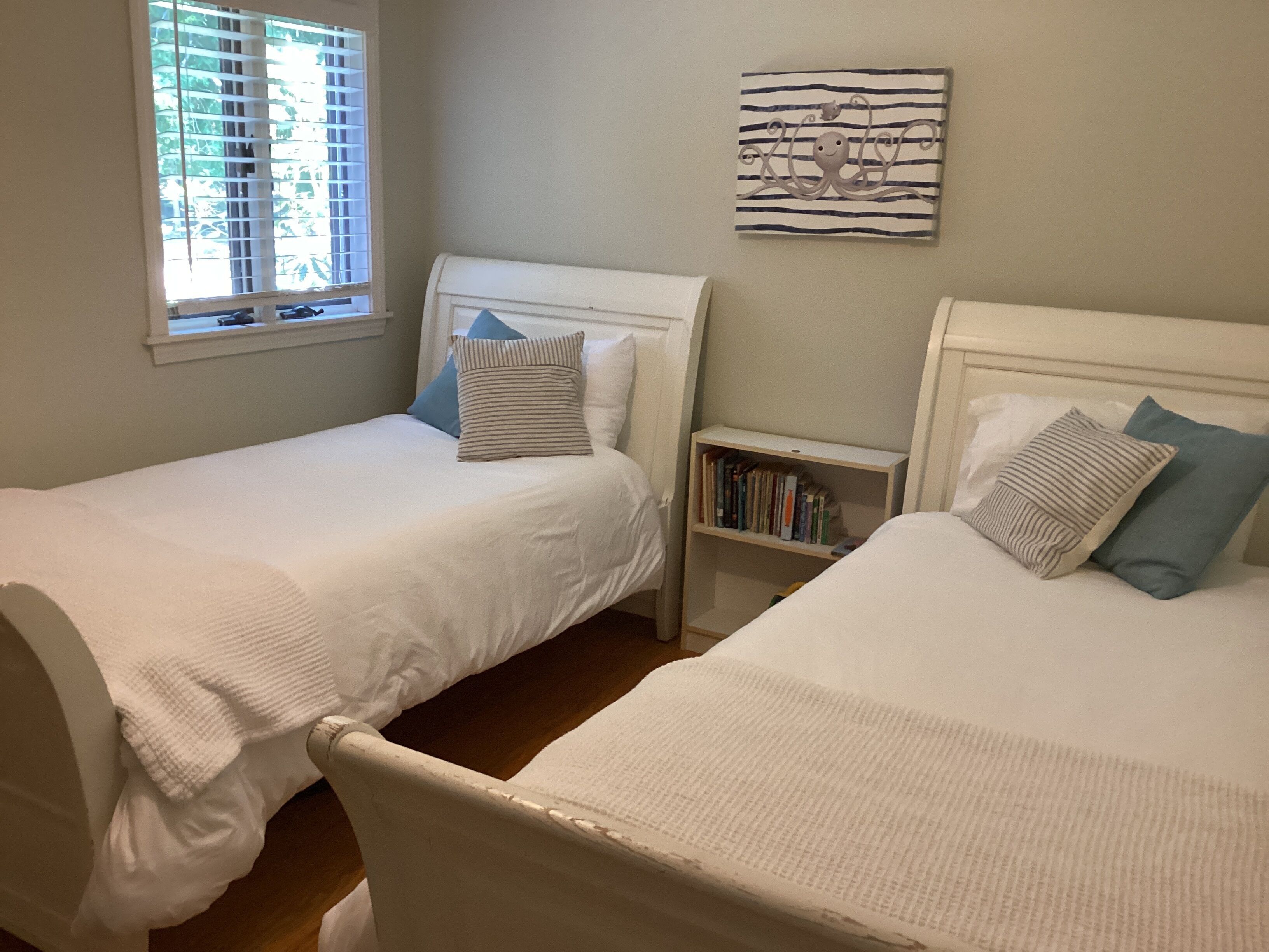 Two twin beds with white bedding, a bookshelf with books, and a window with blinds.