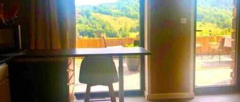 A room with a dining table and chairs, a sofa, and a view of a green landscape through large windows.