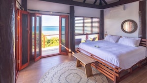 Una camera da letto con un letto grande, pavimento in legno e vista sull'oceano attraverso porte aperte.