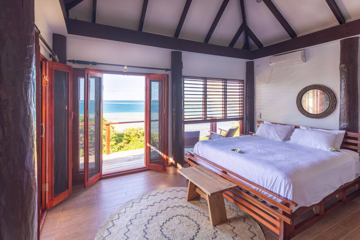 Un dormitorio con una cama grande, piso de madera y vista al océano a través de puertas abiertas.