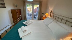Un lit bien fait, avec du linge de lit blanc et deux serviettes, une commode en bois, une lampe et une vue sur l’extérieur par une porte vitrée.