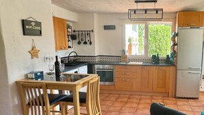 A compact kitchen with wooden cabinets, a dining table with chairs, a refrigerator, and a window with a view of greenery. - La Jacaranda Apartment, Sea Views, Games Room and Private Pool (Mijas)