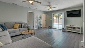 A spacious living room with a grey sofa, a coffee table, and a bookshelf. There are two ceiling fans and a TV mounted on the wall. - Heated Pool Close to St Pete Tampa Pet Friendly (Pinellas Park)