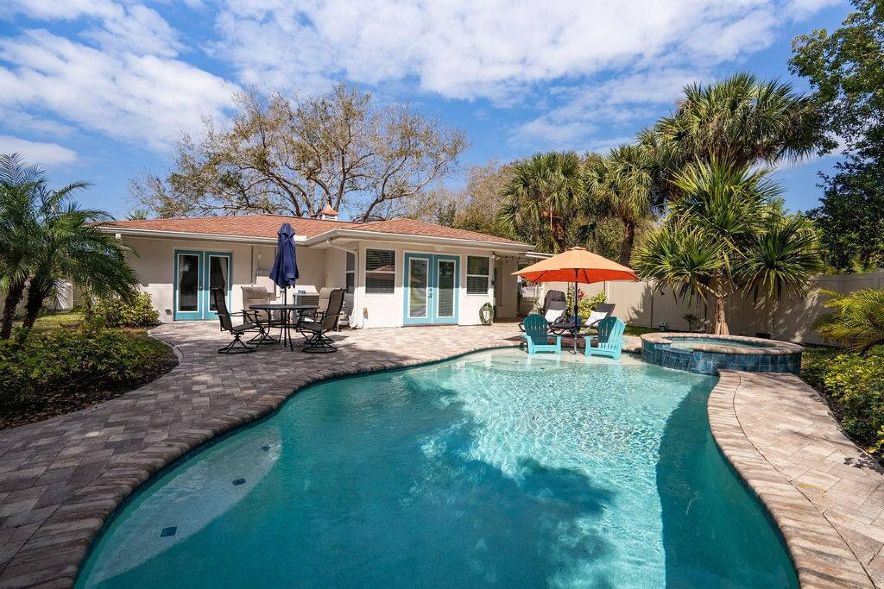 A swimming pool with a curved edge, surrounded by a brick patio. There are lounge chairs and an umbrella near the pool.