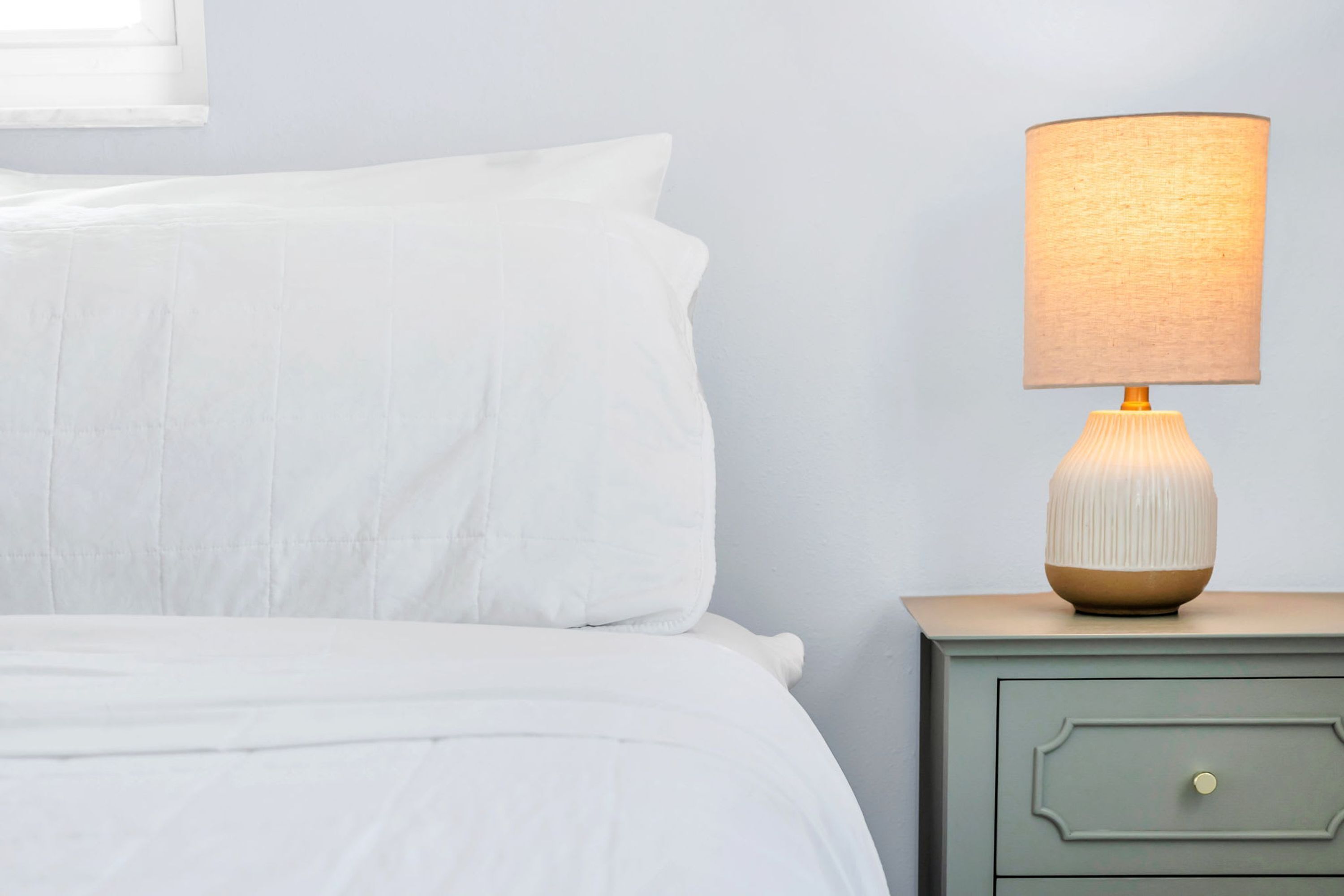 A neatly made bed with white linens, a bedside table with a lamp, and a drawer.