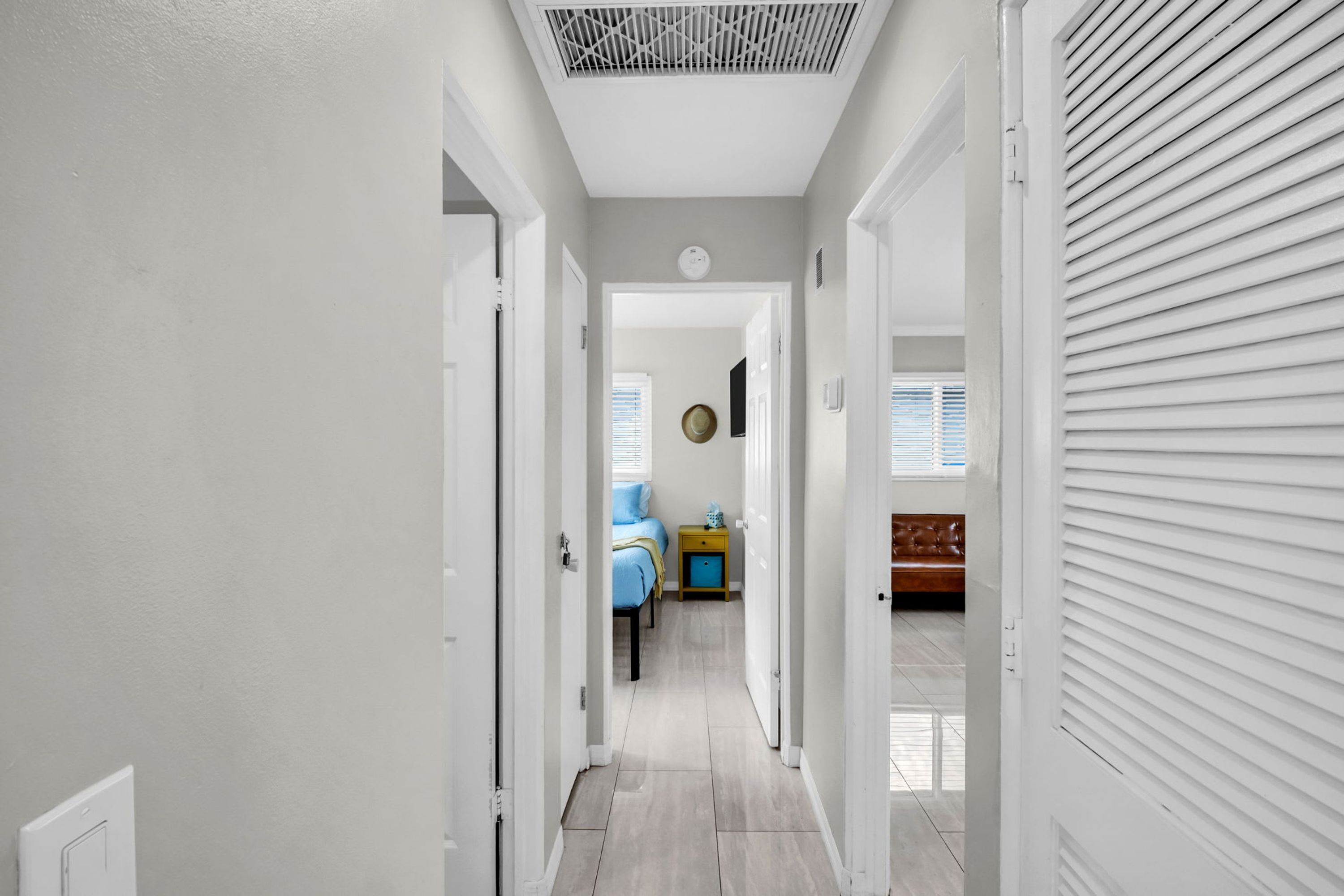 A hallway leading to a bedroom with a bed, bedside table, and a window with blinds.