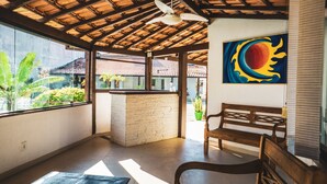 A room with a wooden ceiling, a bench, a wall with a colorful painting, and a view of greenery outside. - Pousada El Camenito (Búzios)