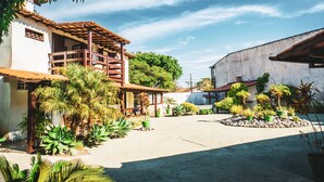 A courtyard with a building featuring a wooden balcony, surrounded by various potted plants and trees. - Pousada El Camenito (Búzios)