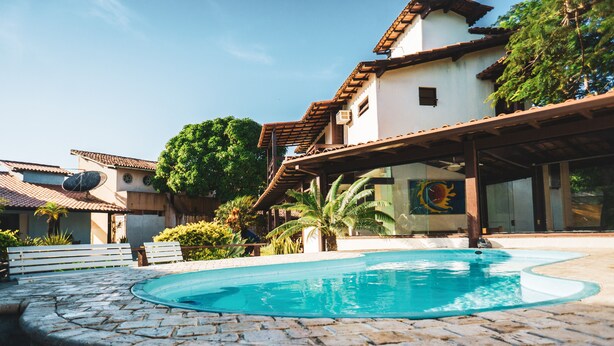 A swimming pool with a stone border, a wooden deck, and a building with a tiled roof and a mural. - Pousada El Camenito (Búzios)