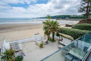 A beach view from a balcony with a wooden deck, outdoor seating, and a glass railing overlooking the sea. - Beachways - 5 Bedroom Holiday Home - Saundersfoot (Saundersfoot)