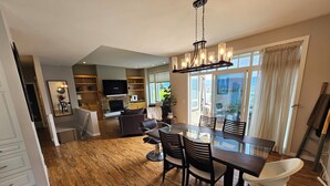 A spacious dining area with a large wooden table and chairs, a modern chandelier, and a view of the outdoors through large windows. - Transcendent View Oasis - Lakeview Family Home (West Kelowna)