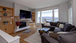 En moderne stue med pejs, et fladskærms-tv, en sofa og store vinduer der giver udsigt til naturen.