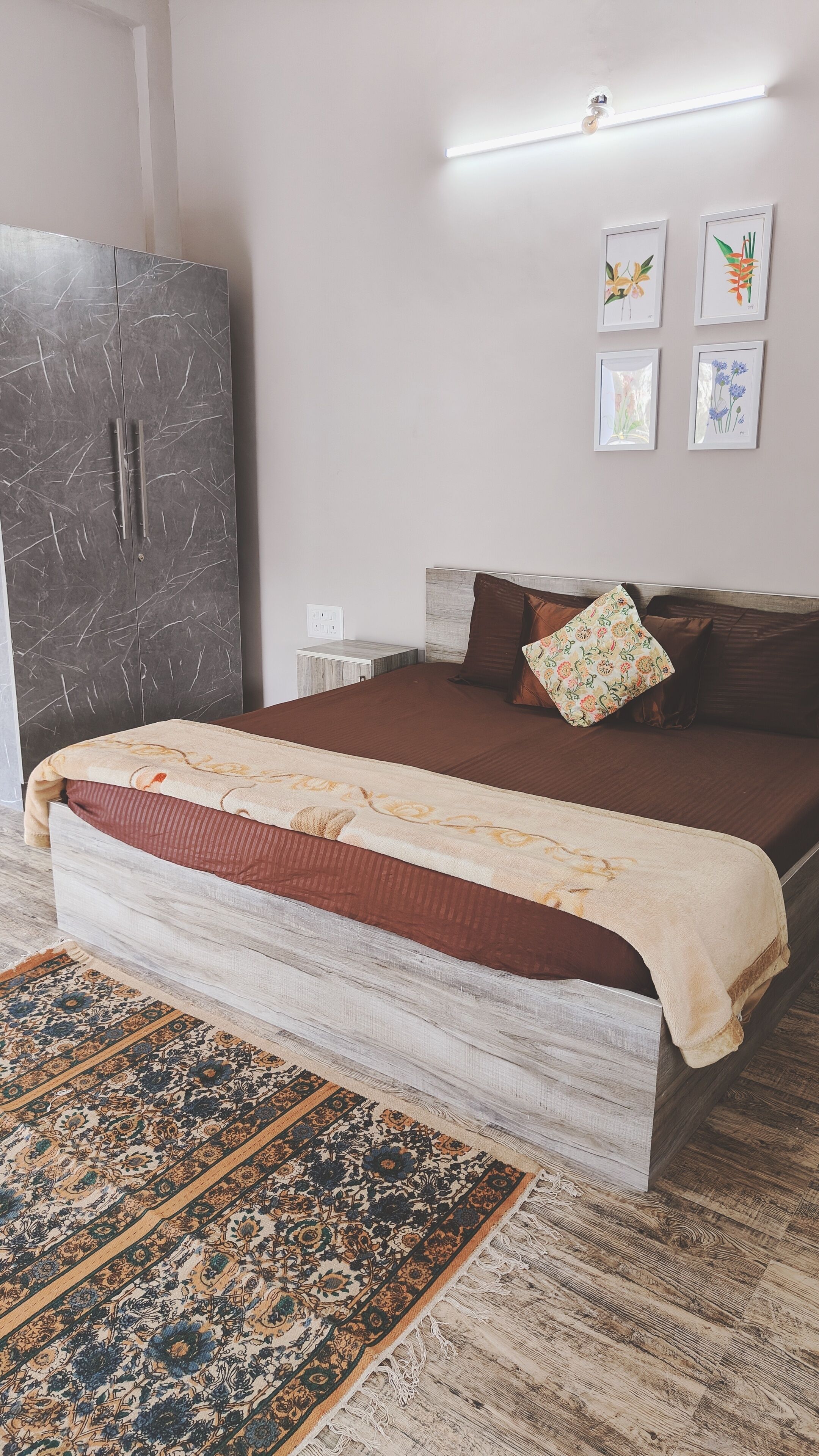 A bedroom with a bed, a wardrobe, a nightstand, a wall-mounted light, and framed artwork.
