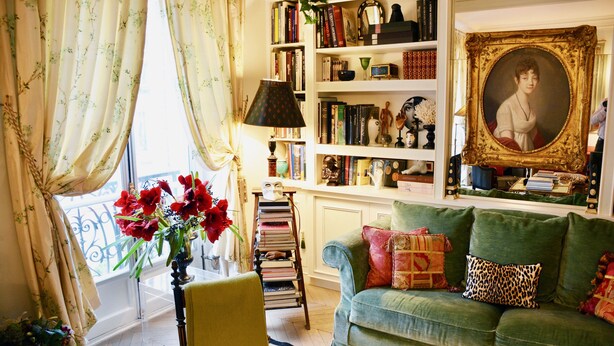 Un salon chaleureux avec un canapé vert, une bibliothèque remplie de livres, un portrait encadré et un vase de fleurs rouges.