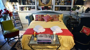 Une chambre élégamment aménagée, dotée d’un grand lit, d’une table de chevet avec des livres et une bougie, d’un fauteuil et d’une bibliothèque remplie de livres et d’objets décoratifs.