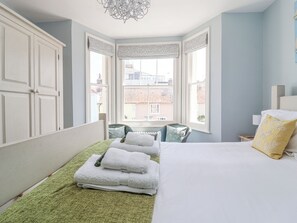 A bedroom with a bed, a chair, a window with curtains, and a wardrobe. - The Corner Cottage (Aldeburgh)