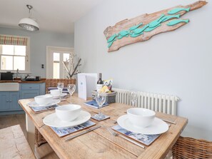 A dining area with a wooden table set for four, white plates, bowls, and wine glasses. A rustic wall art piece and a wicker basket with a bottle and glasses are visible. - The Corner Cottage (Aldeburgh)