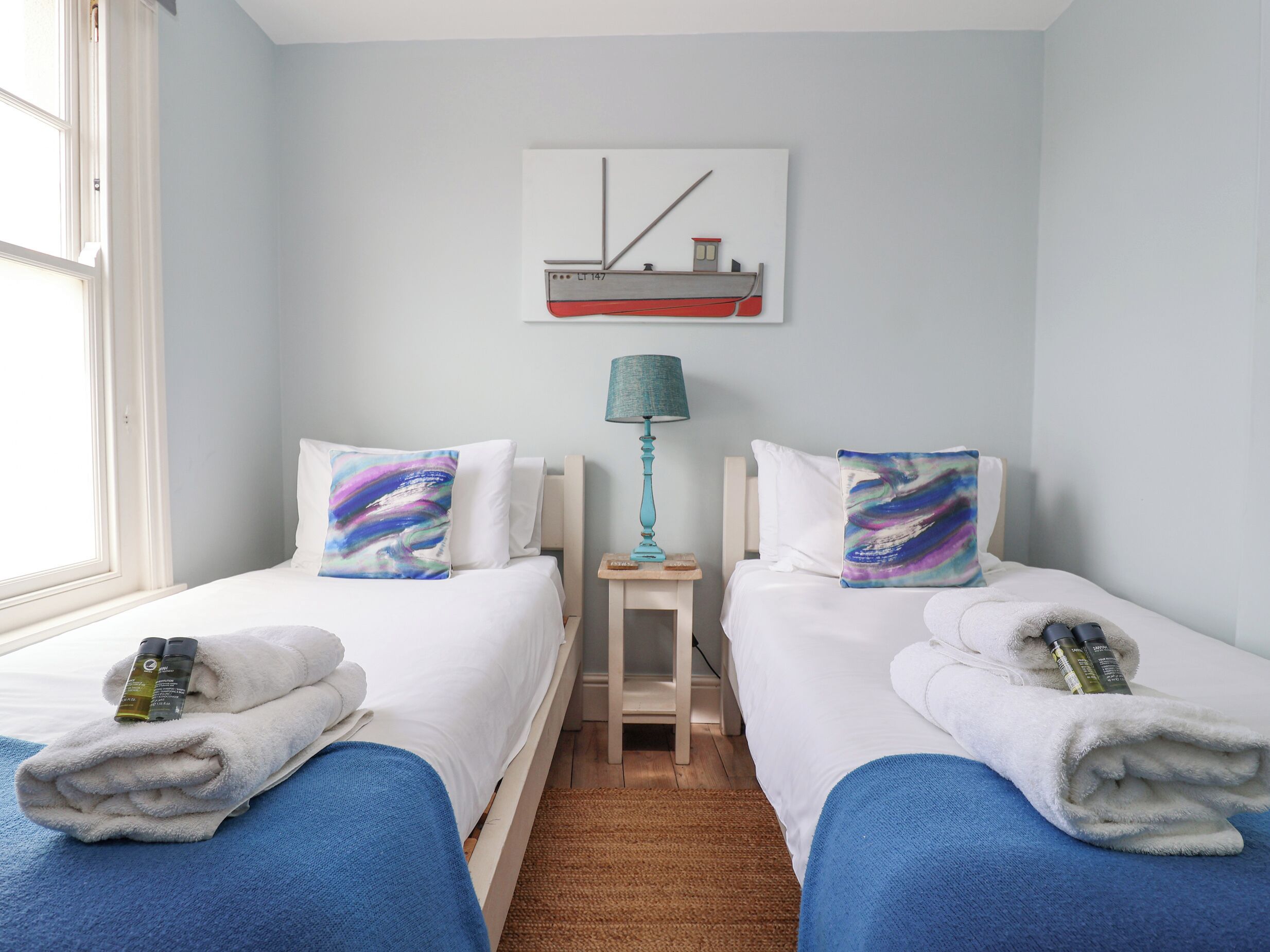 Une chambre d’hôtel avec deux lits, chacun recouvert d’une couverture bleue et agrémenté d’un oreiller orné d’un motif abstrait et coloré. Une petite table avec une lampe se trouve entre les lits, et un tableau représentant un navire est accroché au mur au-dessus de la table.