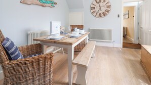 A dining area with a wooden table set for two, a wicker chair, a bench, and a large wall clock. - The Corner Cottage (Aldeburgh)
