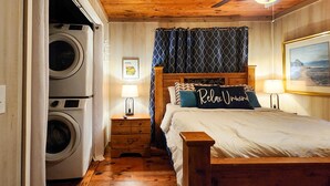 A bedroom with a wooden bed, a nightstand with a lamp, a wall-mounted picture, and a washing machine in the corner. - Bungalow+RV/10 acres/Stream/Sleeps6 (Ellijay)