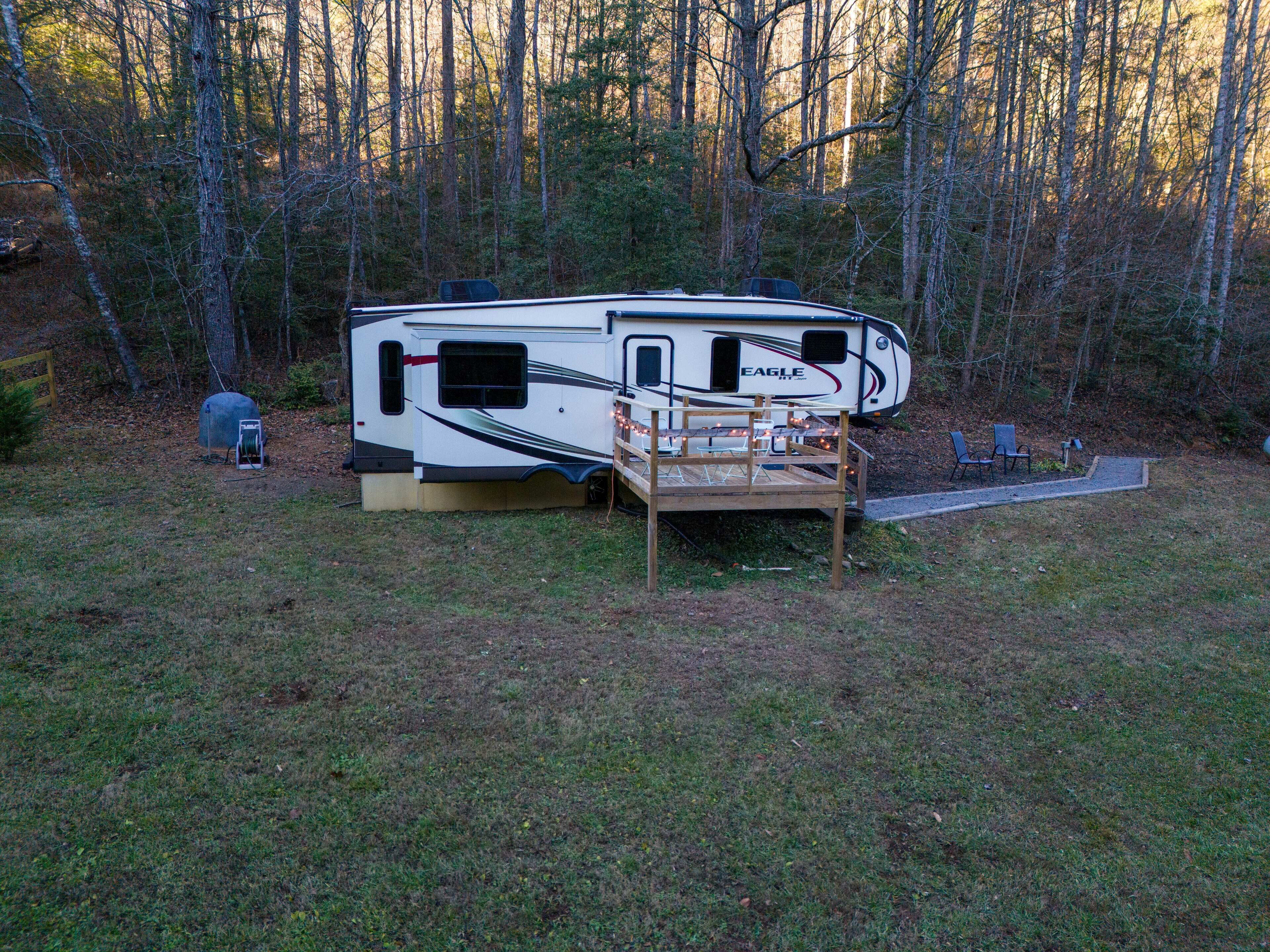 A recreational vehicle (RV) parked in a wooded area with a wooden deck and outdoor seating.