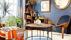 Una sala de estar acogedora con una mesa redonda, un cojín estampado, una silla de madera y una estantería llena de objetos decorativos.