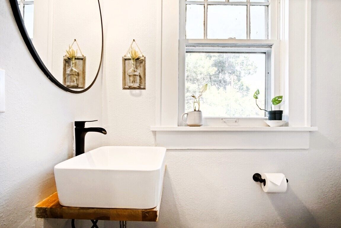 Ein Badezimmer mit einem weißen Waschbecken auf einem Holzboden, einem schwarzen Wasserhahn, einem Fenster mit Blick auf Bäume und zwei Wandleuchten.