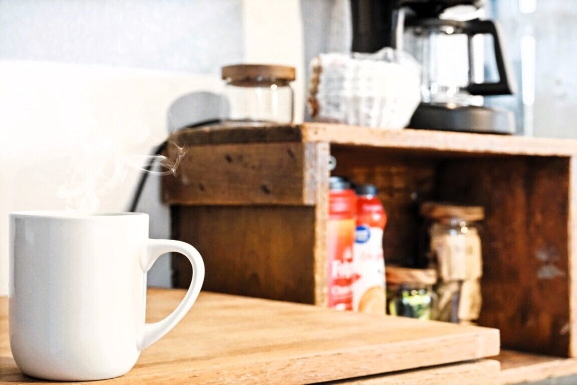 A white mug on a wooden surface with steam rising from it, suggesting hot beverage. In the background, there is a coffee maker and a shelf with jars and containers, indicating a kitchen setting.