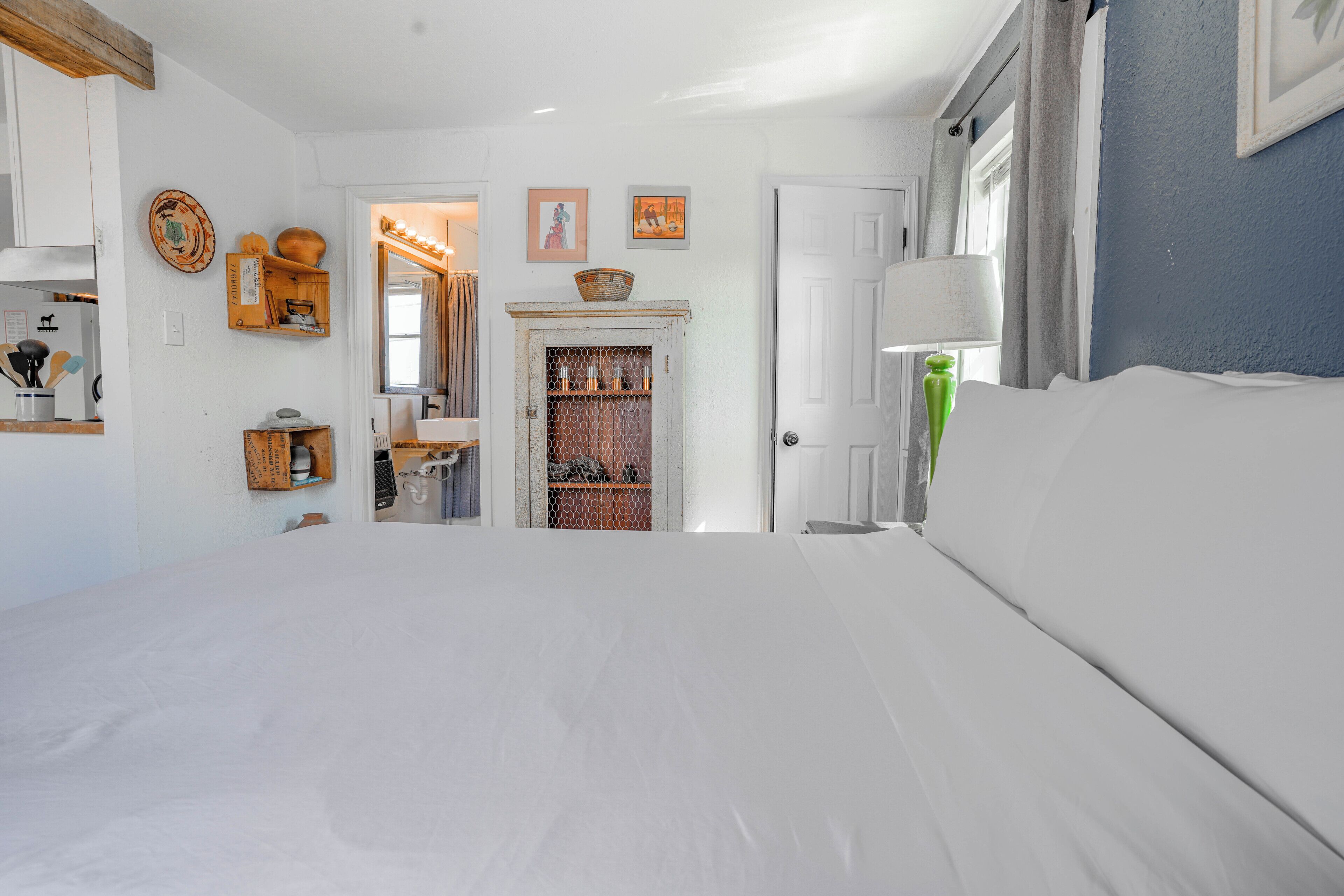 Une chambre à coucher avec un lit, une porte donnant sur une salle de bain et un couloir avec un miroir et des étagères.