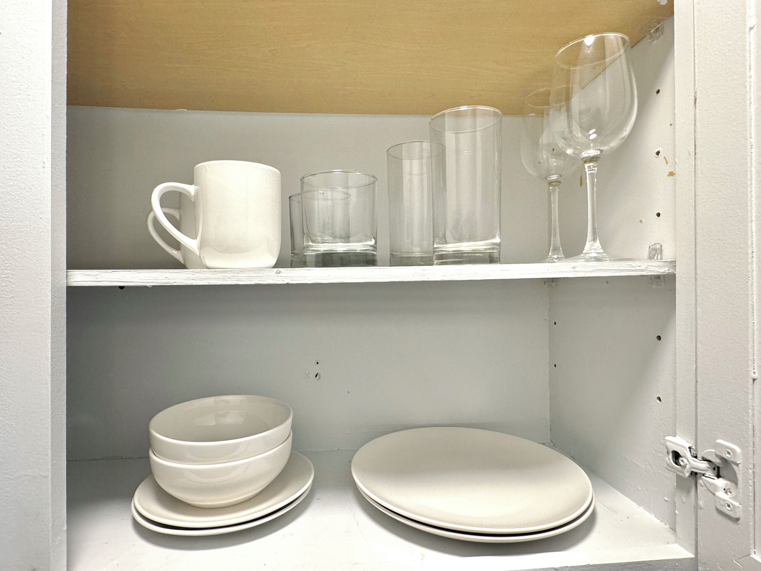 Ein weißes Regal mit klarem Glasgeschirr, darunter eine Tasse, Gläser und ein Weinglas, sowie gestapelte weiße Teller.